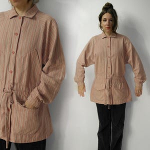 May include: A light pink striped jacket with a collared neckline, button-up front, and two front pockets. The jacket has long sleeves and a drawstring waist. The fabric has vertical stripes in shades of pink and cream.