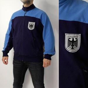 Puede incluir: Chaqueta con cremallera azul marino y azul claro con el texto "BUNDESWEHR" y un escudo. La chaqueta tiene cuello alto, mangas largas y cremallera frontal. Hay un bolsillo a cada lado.