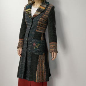 May include: A long, dark gray coat with colorful patterned accents on the collar, sleeves, and pockets. The coat features a button-down front and a flared skirt. The model is wearing a white shirt and red pants.