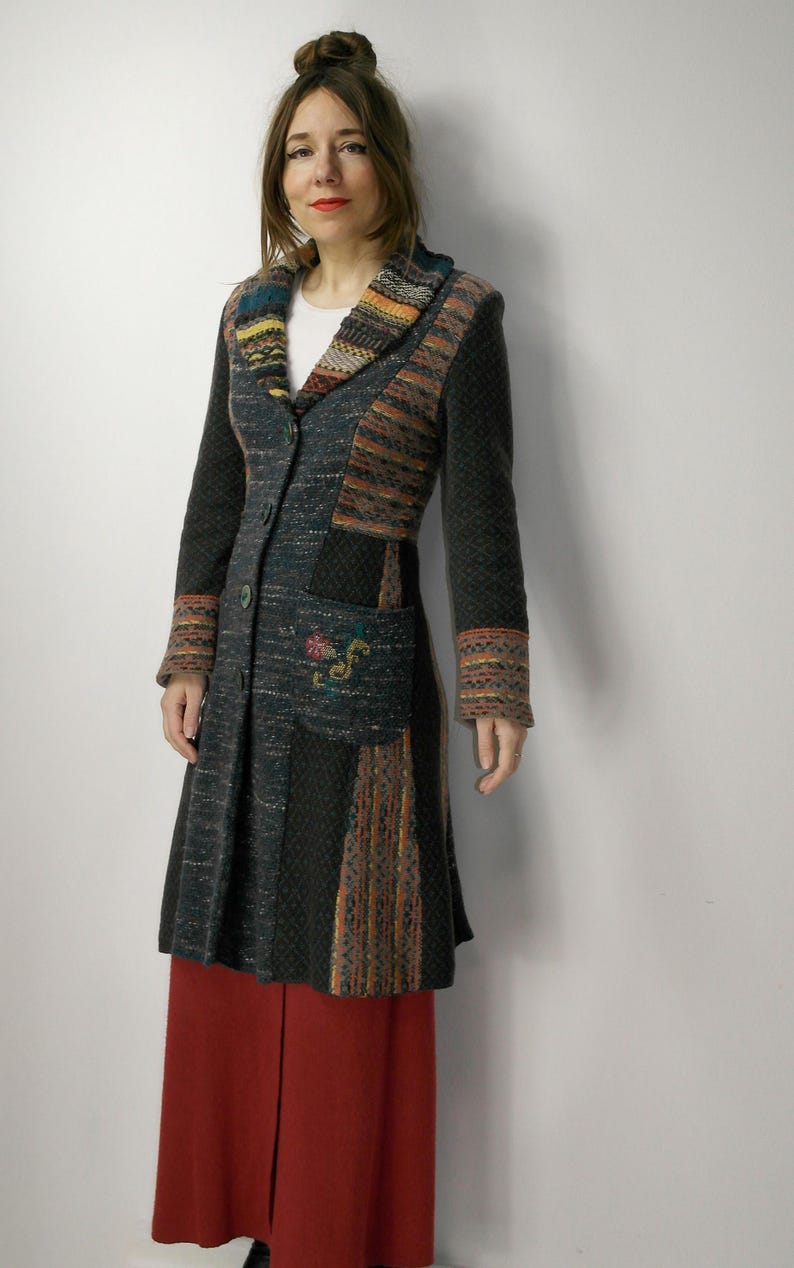 May include: A long, dark gray coat with colorful patterned accents on the collar, sleeves, and pockets. The coat features a button-down front and a flared skirt. The model is wearing a white shirt and red pants.