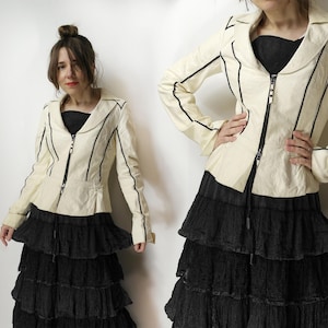 Puede incluir: Chaqueta color crema con ribetes negros y cierre de cremallera. La chaqueta se combina con una falda negra escalonada. El conjunto es elegante y adecuado para diversas ocasiones. La chaqueta presenta un diseño clásico con un toque moderno.