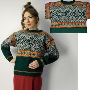 May include: A green, gray, and orange patterned sweater. The sweater features a geometric design across the chest and sleeves, with a solid green lower section. The sleeves and neckline are also green. The sweater is displayed on a person.