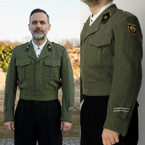 Op de afbeelding: Olijfgroene militaire jas met een hoge kraag, twee borstzakken en een tailleband. De jas heeft schouderinsignes en een armpatch. Het model draagt een wit overhemd en een zwarte broek. De jas heeft lange mouwen met strepen.