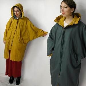 Op de afbeelding: Een omkeerbare, oversized jas in mosterdgeel en blauwgroen. De gele kant heeft een knoopsluiting, terwijl de blauwgroene kant een ritssluiting heeft. Beide kanten hebben een capuchon en grote zakken. De jas is knielang.