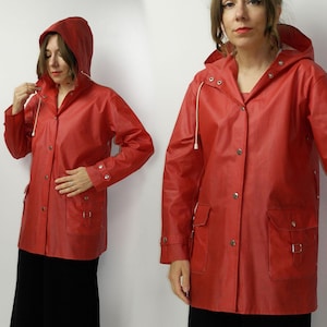 Puede incluir: Un impermeable rojo brillante con capucha, cordón y cierres de botón a presión. La chaqueta tiene dos bolsillos delanteros con solapas y un diseño clásico. El impermeable está hecho de un material brillante e impermeable.