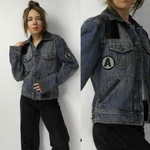 Könnte beinhalten: Jeansjacke in verwaschenem Blau mit schwarzen Patches und Taschen. Die Jacke hat ein klassisches Design mit Knopfleiste und Kragen. Aufnäher mit dem Buchstaben "A" und einem Symbol sind sichtbar.