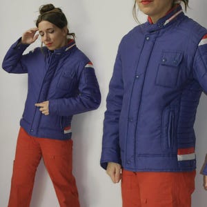 Könnte beinhalten: Eine blaue Vintage-Jacke mit roten und weißen Akzenten an Kragen, Ärmeln und Saum. Die Jacke hat ein gestepptes Design, zwei Vordertaschen und einen Knopfverschluss. Die Jacke wird mit einer orangefarbenen Cargohose kombiniert.
