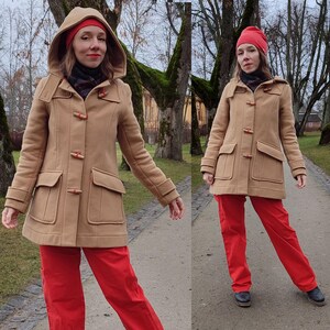 May include: A tan duffle coat with a hood, worn with red trousers and a red beanie. The coat features large pockets and wooden toggles. The person is standing on a paved path with trees in the background.