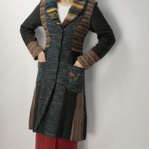 May include: A long, multi-colored coat with a patchwork design. The coat features a mix of textures and patterns, including knitted and woven sections in shades of gray, brown, orange, and blue. It has a collared neckline, green buttons, and pockets with floral embroidery.