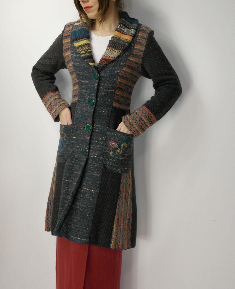 May include: A long, multi-colored coat with a patchwork design. The coat features a mix of textures and patterns, including knitted and woven sections in shades of gray, brown, orange, and blue. It has a collared neckline, green buttons, and pockets with floral embroidery.