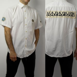 May include: White short-sleeved shirt with a button-down front and collar. The shirt has a logo patch on the chest and sleeve, and a large "NAPAPIJRI Geographic" logo on the back. The shirt is worn with black trousers.
