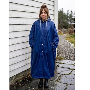 Puede incluir: Un impermeable largo de color azul marino que cuenta con una capucha con cordones blancos, cierre frontal de botones y mangas largas. El abrigo tiene un acabado liso resistente al agua y llega hasta la parte inferior de la pantorrilla.