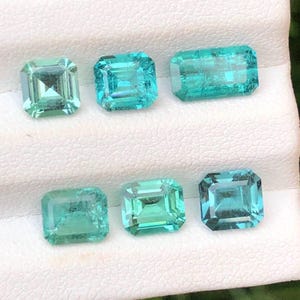 May include: Six faceted gemstones in shades of teal and green are arranged on a white surface. The gemstones are rectangular and square-cut, with visible internal facets. The gems vary slightly in color, from a lighter green to a deeper teal.