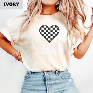 Pu&ograve; includere: T-shirt color avorio con un motivo a cuore a scacchi bianchi e neri. La maglietta &egrave; di colore crema chiaro e ha maniche corte. La parola "IVORY" &egrave; stampata in nero in grassetto nell'angolo in alto a sinistra dell'immagine.