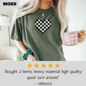 Pu&ograve; includere: T-shirt verde oliva con un cuore a scacchi bianco e nero. La maglietta ha una vestibilit&agrave; ampia e maniche corte. La parola "MOSS" &egrave; nell'angolo in alto a sinistra. Una tazza rosa con le parole "Cool Mom Club" &egrave; tenuta in mano.