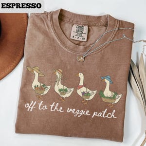 May include: Espresso-colored t-shirt with a graphic of four geese wearing hats and carrying baskets of vegetables. The text "off to the veggie patch" is printed below the geese. A silver necklace is draped across the shirt.