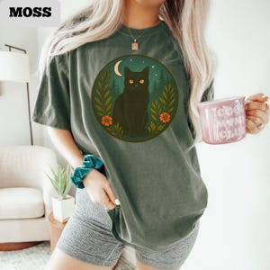May include: A moss green t-shirt featuring a circular graphic of a black cat with yellow eyes, a crescent moon, and foliage. The word "MOSS" is printed in the upper left corner. The person is holding a pink mug with the words "Cool Mom Club".