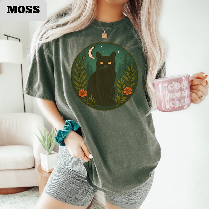 May include: A moss green t-shirt featuring a circular graphic of a black cat with yellow eyes, a crescent moon, and foliage. The word "MOSS" is printed in the upper left corner. The person is holding a pink mug with the words "Cool Mom Club".