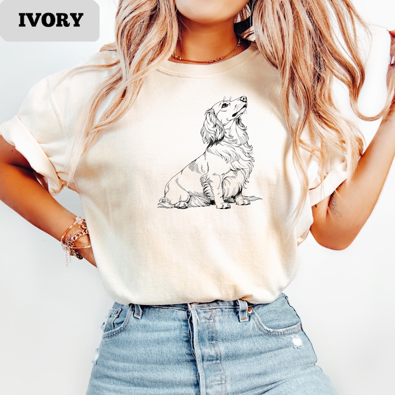 May include: Ivory-colored t-shirt featuring a detailed black and white illustration of a long-haired dachshund dog looking upwards. The word "IVORY" is printed in a bold font in the upper left corner. The shirt is made of a soft material.