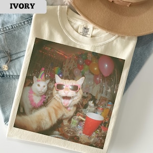 May include: Ivory-colored t-shirt featuring a colorful image of cats at a party. One cat wears sunglasses and another a party hat. The shirt is made of a soft material and has a Comfort Colors tag.