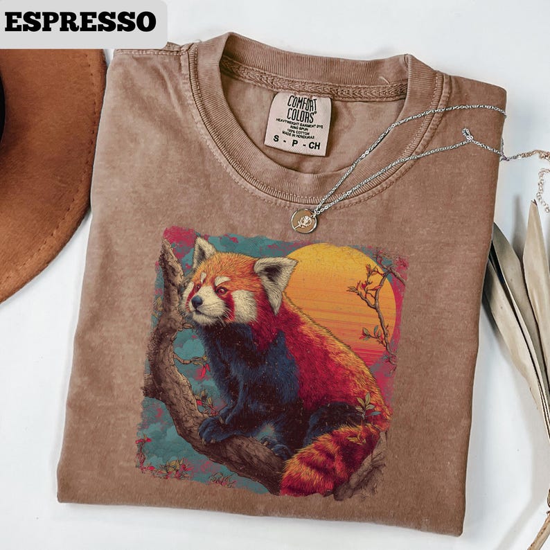 Puede incluir: Camiseta color espresso con un gr&aacute;fico de un panda rojo en la rama de un &aacute;rbol con un fondo de puesta de sol. La camiseta tiene cuello redondo y mangas cortas. Un collar plateado con un colgante est&aacute; sobre la camiseta.
