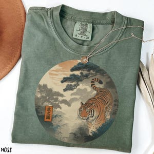 May include: A moss green crew neck t-shirt featuring a circular graphic print of a tiger walking through a misty forest landscape with pine trees and a sun. A rectangular orange label with black text reading 虎之圖 is visible on the left side of the print.