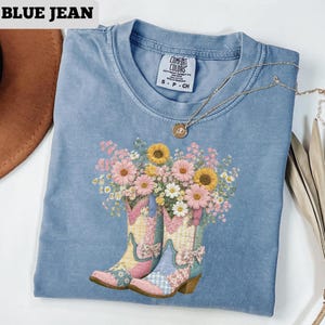 May include: Blue jean-colored t-shirt featuring a floral design of cowboy boots filled with sunflowers and daisies. The shirt has a crew neck and short sleeves. A gold necklace with a pendant is draped across the shirt.