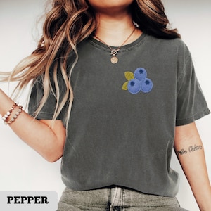 May include: A gray t-shirt with a small embroidered design of three blue blueberries and two green leaves. The word "PEPPER" is printed in a white, bold font at the bottom of the image.