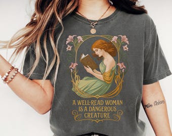 May include: A charcoal gray t-shirt featuring a graphic of a woman with auburn hair reading a book, framed by pink lilies and gold vines. Below the illustration, the text reads: A WELL-READ WOMAN IS A DANGEROUS CREATURE in gold lettering.