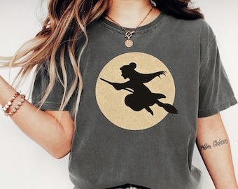 May include: Pepper-colored t-shirt featuring a black silhouette of a witch flying on a broom in front of a tan-colored full moon. The shirt has a crew neck and short sleeves. The word "PEPPER" is printed in a white, bold font at the bottom.