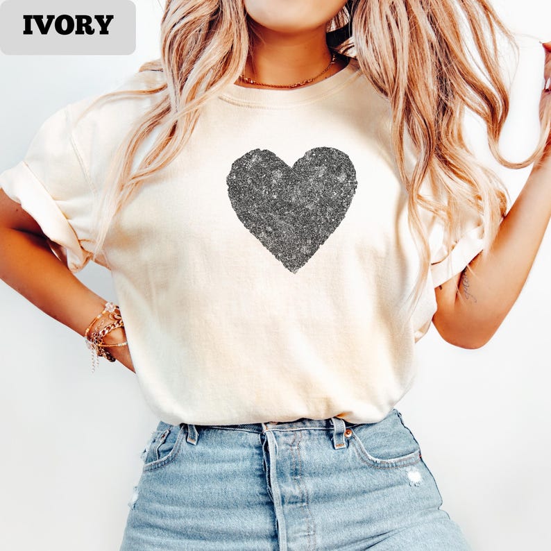 K&ouml;nnte beinhalten: Elfenbeinfarbenes T-Shirt mit einem gro&szlig;en, glitzernden Herzdesign. Das Herz ist mittig auf dem Shirt platziert. Das Shirt hat kurze &Auml;rmel und eine lockere Passform. Das Wort "IVORY" ist in einem rechteckigen Feld oben links aufgedruckt.