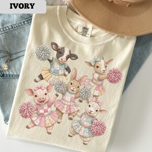May include: Ivory-colored t-shirt featuring a graphic of farm animals dressed as cheerleaders. The animals include a cow, pig, rabbit, and goats, each holding pom-poms and wearing skirts. The shirt has a Comfort Colors tag.
