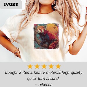 Puede incluir: Camiseta de color marfil con un gr&aacute;fico vibrante de un panda rojo posado en una rama contra un fondo de puesta de sol. La camiseta tiene cuello redondo y mangas cortas. La palabra "IVORY" est&aacute; impresa en la parte superior.