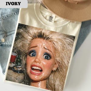 May include: Ivory-colored t-shirt featuring a distressed image of a doll with wide blue eyes and an open mouth. The shirt has a Comfort Colors tag. A tan hat and blue jeans are partially visible in the background.