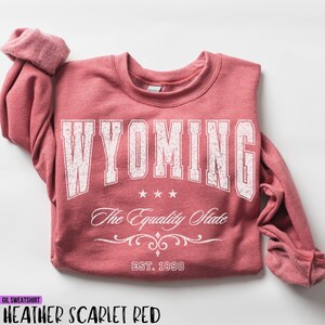 May include: A heather scarlet red crewneck sweatshirt featuring the white text WYOMING in a distressed block font, three small stars, the script text The Equality State, a decorative flourish, and the text EST. 1890 at the bottom.