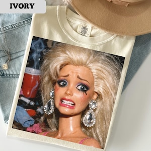 May include: Ivory-colored t-shirt with a graphic of a crying doll with blonde hair, wearing large diamond earrings. The shirt has a Comfort Colors tag. The word "IVORY" is printed in a white box in the upper left corner.