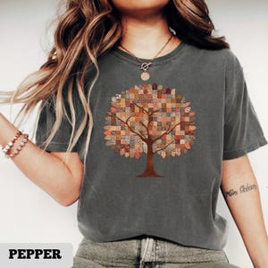 Puede incluir: Camiseta gris oscuro con un diseño de árbol. El dosel del árbol está formado por un patrón de patchwork. El tronco y las ramas son marrones. La palabra "PEPPER" está impresa en la parte inferior de la camiseta.