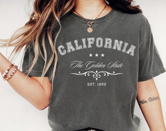 May include: Pepper-colored t-shirt with the word "CALIFORNIA" in white, arched across the top. Below, it reads "The Golden State" with three stars and decorative flourishes. The text "EST. 1950" is at the bottom.
