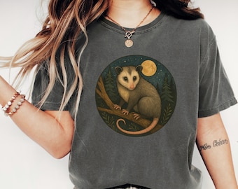May include: Dark gray t-shirt with a circular graphic featuring an opossum on a branch, set against a forest and moon backdrop. The word "PEPPER" is printed at the bottom of the shirt.