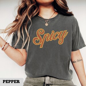 May include: Dark grey t-shirt with the word "Spicy" in a retro orange and gold design. The shirt is a short-sleeved, crew neck style. The word "Pepper" is at the bottom of the image.