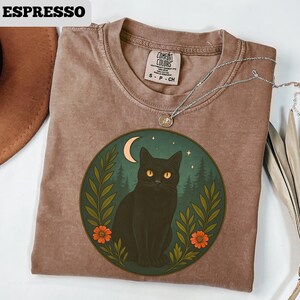 May include: Espresso-colored t-shirt with a round graphic featuring a black cat with yellow eyes, a crescent moon, and a forest scene. The graphic is framed by green leaves and orange flowers. A silver necklace is draped across the shirt.