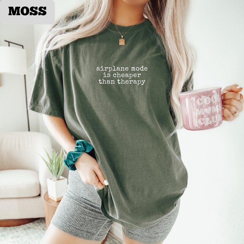 May include: An olive green t-shirt with the text "airplane mode is cheaper than therapy" in white. The person is wearing gray shorts and holding a pink mug with the words "cool mom club" on it. A teal scrunchie is on the wrist.