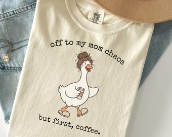 May include: Ivory-colored t-shirt with the text "off to my mom chaos but first, coffee." A cartoon goose with a messy bun and coffee cup is printed on the front. The shirt is made of a soft material and is paired with jeans and a hat.