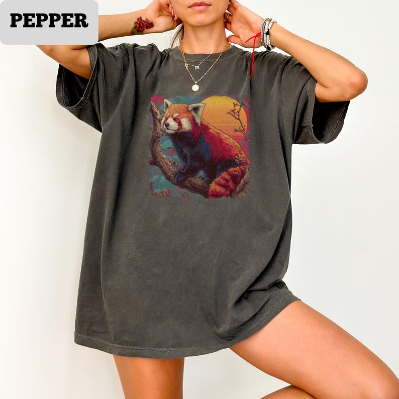 Puede incluir: Camiseta gris oscuro con un gr&aacute;fico colorido de un panda rojo posado en una rama contra un fondo de puesta de sol. La palabra "PEPPER" est&aacute; en un rect&aacute;ngulo blanco. La camiseta tiene mangas cortas y un corte holgado. El dise&ntilde;o es de inspiraci&oacute;n vintage.