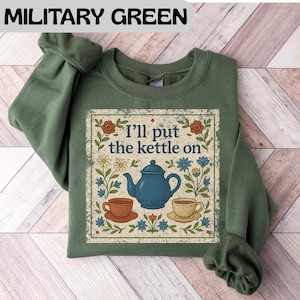 May include: Military green sweatshirt with a graphic that reads "I'll put the kettle on." The graphic features a blue teapot, two teacups, and floral accents. The sweatshirt is a solid color with long sleeves.
