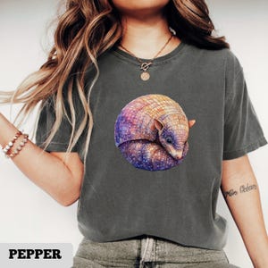 May include: A dark grey t-shirt featuring a watercolor illustration of a curled-up armadillo. The armadillo is depicted with a colorful shell in shades of purple, orange, and yellow, with a starry night sky design. The word "PEPPER" is at the bottom.