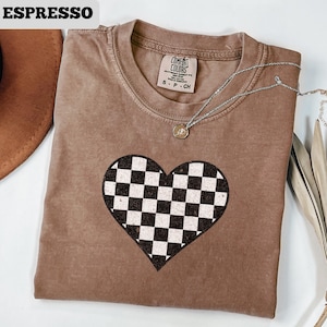 Pu&ograve; includere: T-shirt color espresso con un design a cuore a scacchi. La maglietta ha un girocollo e maniche corte. Una collana d'argento con un piccolo ciondolo &egrave; drappeggiata sulla maglietta. La maglietta &egrave; realizzata in un materiale morbido e confortevole.