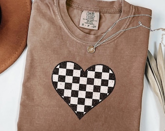 May include: Espresso-colored t-shirt with a checkered heart design. The shirt has a crew neck and short sleeves. A silver necklace with a small pendant is draped on the shirt. The shirt is made of a soft, comfortable material.