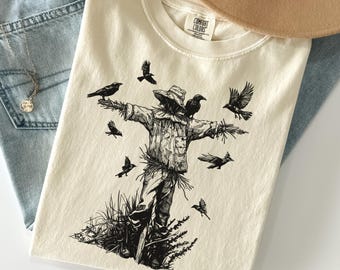 May include: An ivory-colored t-shirt featuring a black ink-style graphic of a scarecrow standing in a field, surrounded by several flying and perched crows. The shirt has a crew neckline and a Comfort Colors brand label printed on the inner collar.