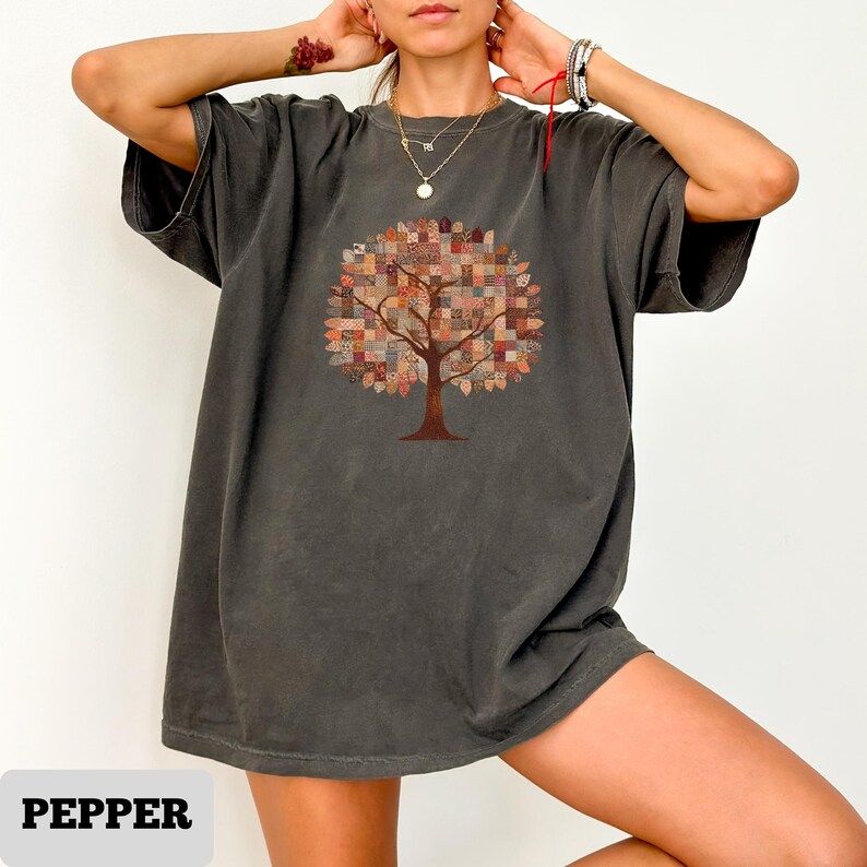 May include: Dark grey t-shirt featuring a tree design with a patchwork-style canopy in shades of brown, orange, and beige. The word "PEPPER" is printed at the bottom of the shirt in a white rectangular box. The shirt has short sleeves and a relaxed fit.